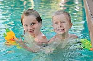 Two smiling siblings in a pool standing with a water pistols