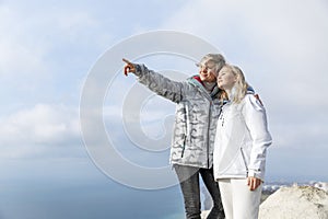 Two smiling women in sports jackets stand on a mountain top above the sea. Active lifestyle, energy and health