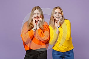 Two smiling young blonde twins sisters girls in vivid colorful clothes putting hands on cheeks isolated on pastel violet