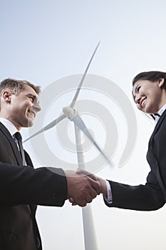 Two smiling young business people shaking hands in front of a wind turbine