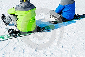 Two snowboarders are sitting on the snow at the top of the mountain and looking out into the distance. View from behind