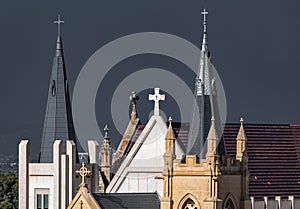 Two spires with crosses and crucifixs on ornate church roof with turrets bathed in sunlight deep grey stormy smokey sky