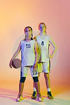 Two sportive girls, teens, basketball players standing together with basketball ball isolated on pink background in neon