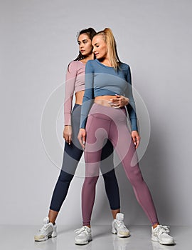Two sporty blonde and brunette girls in athletic body cloth sport wear cloth stand together after workout on gray