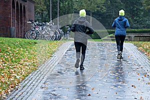 Two sporty female people from behind with neon yellow caps jogging in the rain through an autumn park, there is no bad weather for