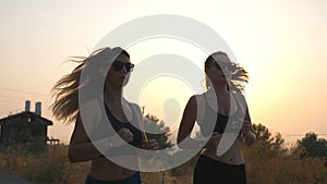 Two sporty women jogging along country road with sunset at background. Young girls working out outdoor at evening time