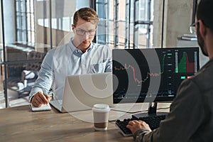 Two stock traders co-working at office sitting at table in front of computers opposite each other