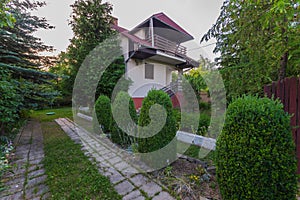 Two-storey white building with red socle and roof. Green bushes grow around it