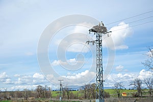 Two storks stand in a nest that is located on top of an electric tower against a blue sky with clouds.