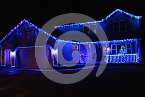 A two story house decorated with blue Christmas lights on the roofline and front porch with a lit Christmas tree in the window