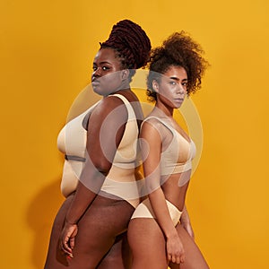 Two strong african american women in beige underwear with different body size standing together back to back, posing