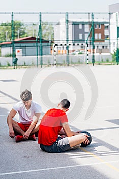 Two strong caucasian athletes resting on the ground at the basketball court while having a conversation