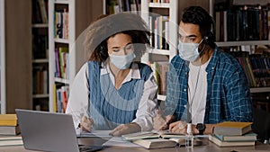 Two students in medical masks sit at desk in university library listen to teacher in headphones on laptop study write