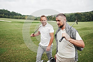 Two stylish men holding bags with clubs and walking on golf course