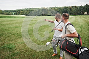 Two stylish men holding bags with clubs and walking on golf course