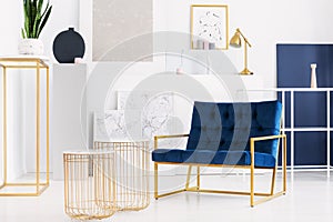 Two stylish tables next to petrol blue armchair in bright living room interior of modern apartment