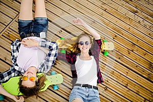 Two stylish young urban girls with longboards lie on the wooden flooring in the street. Friends have fun and spend time