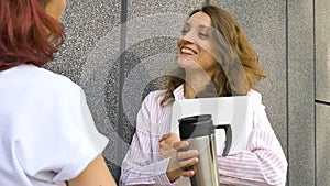 Two successful businesswomen are talking about a the project while standing near the wall outside with a laptop during a