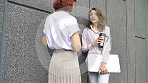 Two successful businesswomen are talking about a the project while standing near the wall outside with a laptop during a