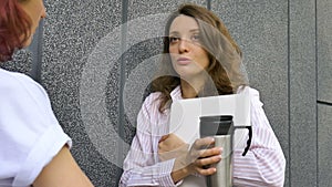 Two successful businesswomen are talking about a the project while standing near the wall outside with a laptop during a