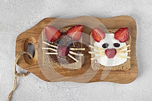 Two sweet toasts for kids in shape of cat with strawberries, banana, cream cheese and chocolate on wooden board