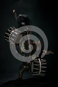 Two taiko drummers in a wig and a demon mask on stage with drumsticks and drums acrobatic stand on a black background.