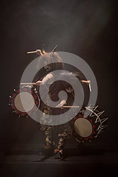 Two taiko drummers in a wig and a demon mask on stage with drumsticks and drums on a black background in light rays.