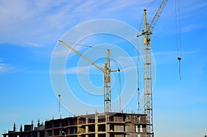 Two tall crane stand near a house under construction on blue sky background.