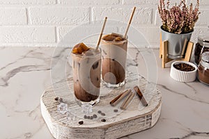 Two tall glass with a chocolate drink or cold coffee with milk and ice cubes stand on a wooden podium opposite a white brick wall