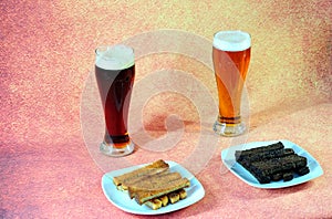 Two tall glasses with light and dark beer stand in front of two plates of fresh wheat and rye croutons on a brown background