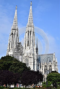 Two tall spiers of a large Gothic church rise above the trees in the blue sky spotted with white clouds.