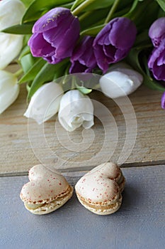 Two tasty creamy macarons are lying on the wooden table with white and violet tulips. Two macarons in form of the heart as 8