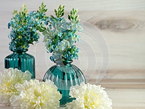 Two teal glass vases up close with blue hydrangeas flowers and white carnations in front of them on a beachwood background.  Home
