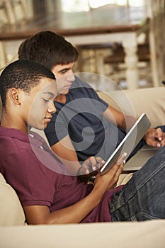 Two Teenage Boys Sitting On Sofa At Home Using Tablet Computer And Laptop