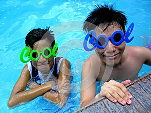 Two teenage boys wearing sunglasses with the word cool for its frame in a swimming pool