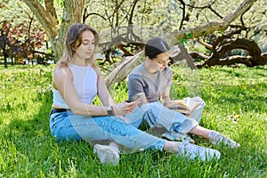 Two teenage girls friends relaxing on grass using smartphone reading book