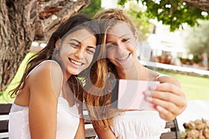 Two Teenage Girls Sitting On Bench Taking Selfie In Park