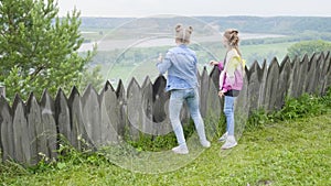 Two teenage girls stand by the wooden fence looking into distance. Girls admiring summer landscape forest and river from