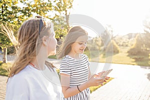 Two teenage girls are walking down the street and chatting in the summertime. A cute girl in casual clothes shows a smartphone to