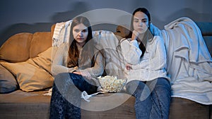 Two teenage girls watching TV on sofa and eating popcorn at night