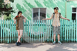 Two teenagers boy and girl going down the street to school, they posing at the green fence near the house, education and back to