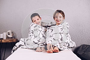 Two teenagers are laughing wrapped in a blanket. A girl and a boy are sitting on the bed hugging each other. Happy brother