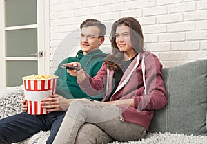 Two teenagers are sitting on the couch watching TV