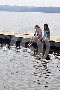 Two teens sitting in bathing suits on wooden dock with legs dangling in lake water with copy space