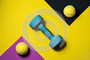 Two tennis balls and a blue dumbbell on a lilac, black and yellow background. Sport, activity, fitness