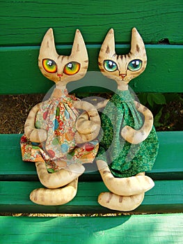 Two textile toy cats with big expressive eyes are sitting on a green bench on a sunny summer day.