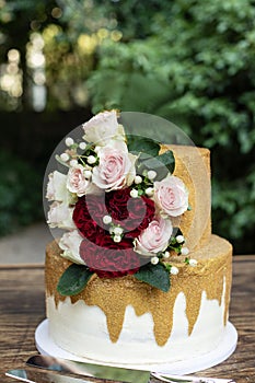 Two tiered white and gold wedding cake with red and pink roses and ruscus leaves