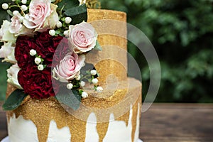 Two tiered white and gold wedding cake with red and pink roses and ruscus leaves