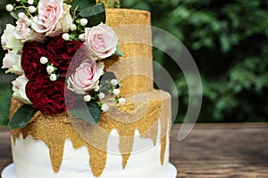 Two tiered white and gold wedding cake with red and pink roses and ruscus leaves