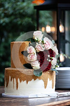 Two tiered white and gold wedding cake with red and pink roses and ruscus leaves